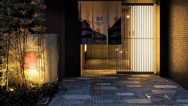 Hotellin sisäänkäynti, jossa on «MUSUBI HOTEL» -tekstillä varustettu banneri, sekä perinteinen japanilainen puutarha, jossa on askelpolkuja ja bambua.