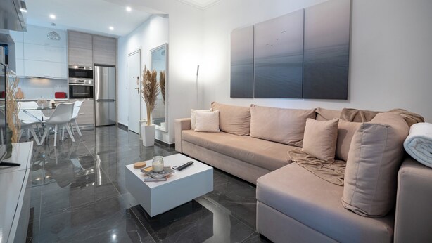 A modern living room with a beige sofa, a coffee table, and a dining area with a glass table and chairs. - Neo Irakleio train st 1 bedrm 4 pers apartment by MPS (Iraklio)