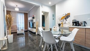 A modern living room with a glass dining table, white chairs, and a sofa. There is a kitchen area with a coffee machine and cabinets. - Neo Irakleio train st 1 bedrm 4 pers apartment by MPS (Iraklio)