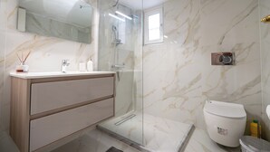 A modern bathroom with a glass-enclosed shower, a marble wall, a large mirror, and a toilet with a trash bin. - Neo Irakleio train st 1 bedrm 4 pers apartment by MPS (Iraklio)
