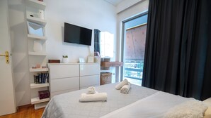 A hotel room with a bed, a TV mounted on the wall, a window with curtains, and a balcony with a view. - Neo Irakleio train st 1 bedrm 4 pers apartment by MPS (Iraklio)