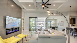 A modern living room with a large flat-screen TV, a ceiling fan, and a dining area with a bar. - Villa Suntastic - gulf access, pool & spa (Cape Coral)