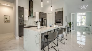 A modern kitchen with a marble island, bar stools, stainless steel appliances, pendant lighting, and a large window. - Villa Suntastic - gulf access, pool & spa (Cape Coral)
