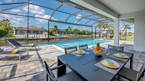 A poolside dining area with a table set for two, lounge chairs, and a view of a swimming pool and palm trees. - Villa Suntastic - gulf access, pool & spa (Cape Coral)