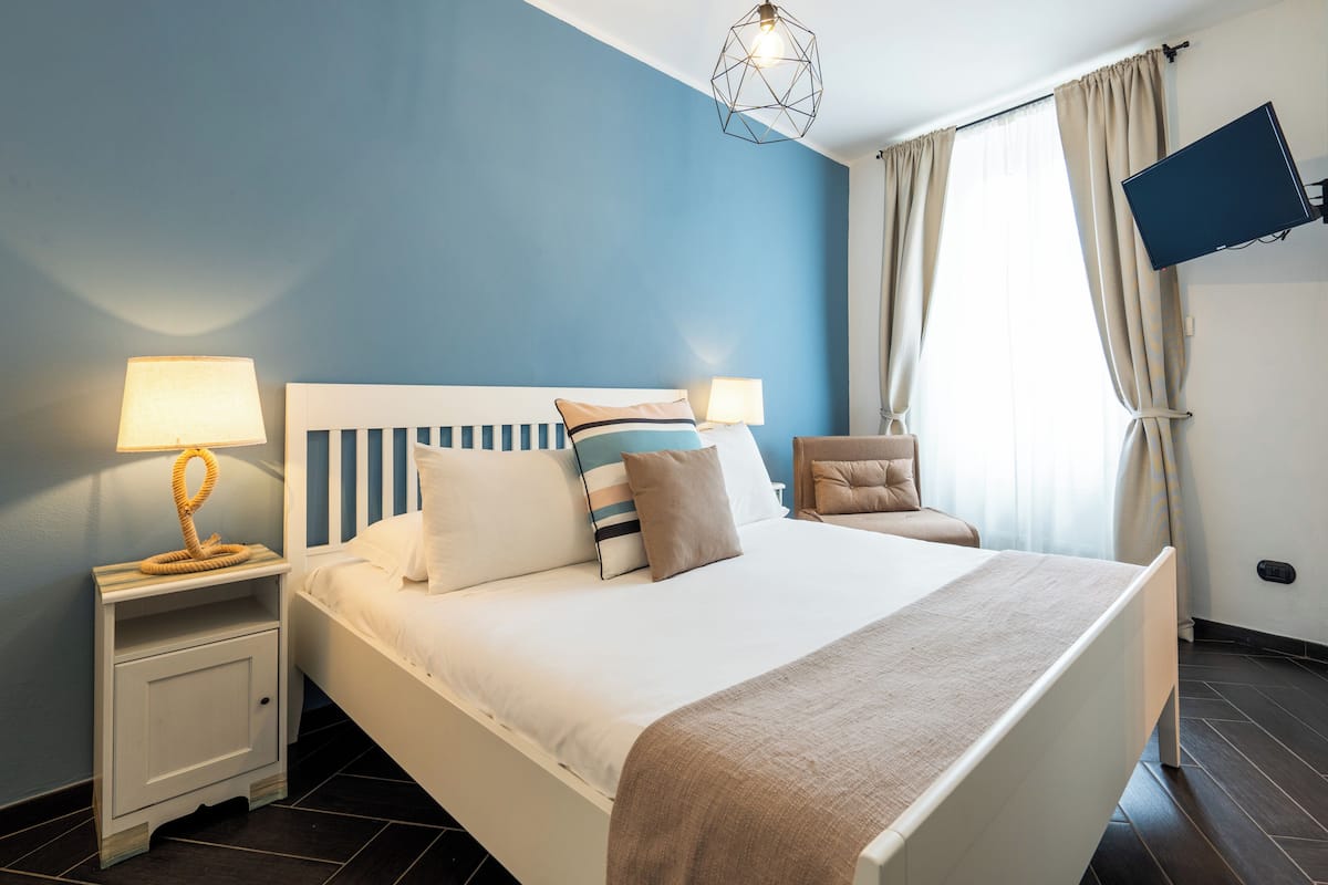 Ein ordentlich eingerichtetes Schlafzimmer mit einem Bett, Nachttischlampen, einem an der Wand montierten Fernseher und einem Fenster mit Vorhängen.