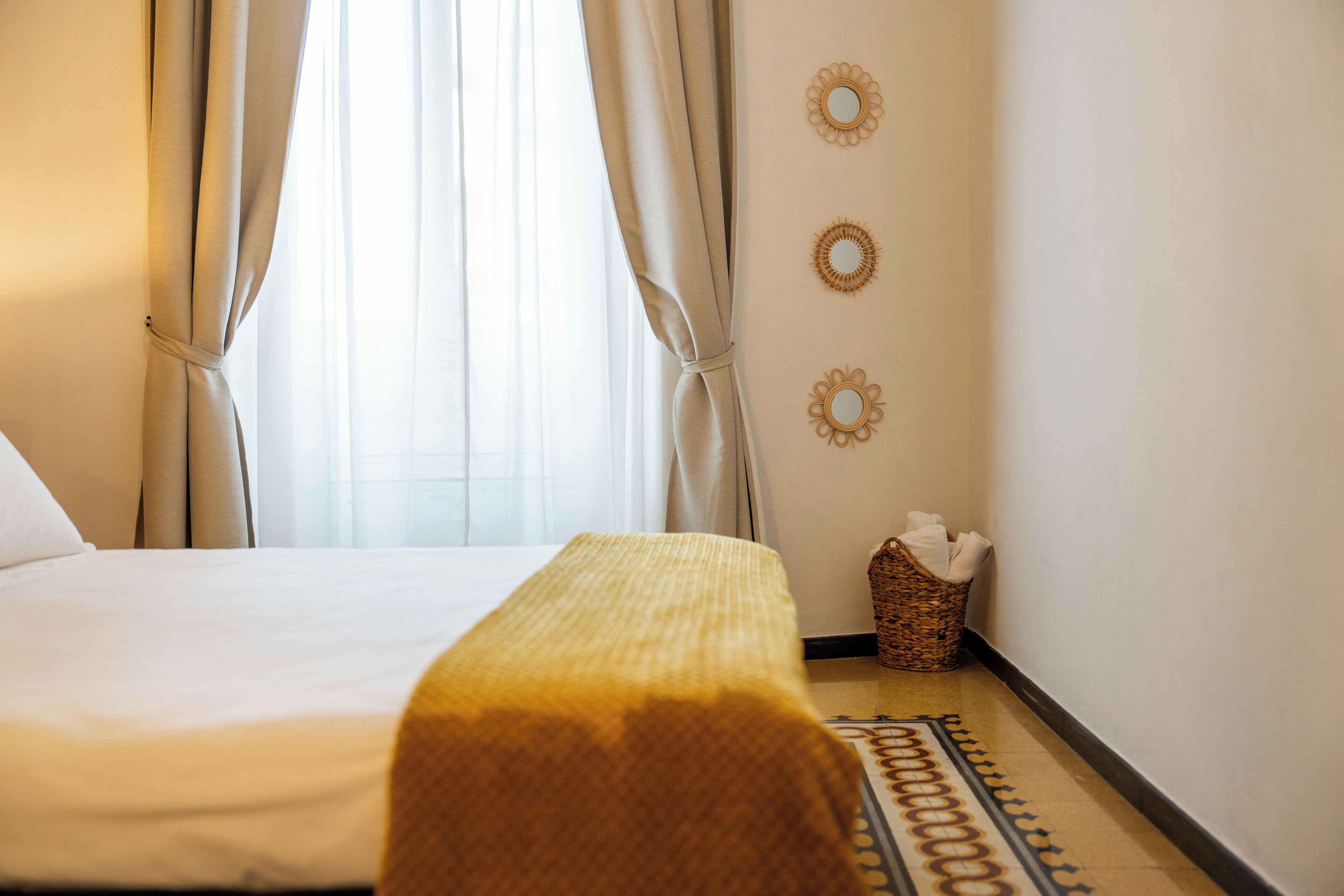 Habitación de hotel con cama, cortinas, cesta de noche y espejos decorativos en la pared.