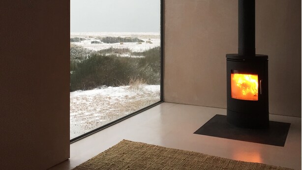 Een modern interieur met een groot raam dat uitzicht biedt op een besneeerd landschap, een zwarte houtkachel en een minimalistisch ontwerp.
