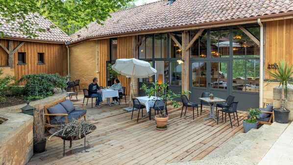 An outdoor patio with wooden tables and chairs, a white umbrella, and potted plants.