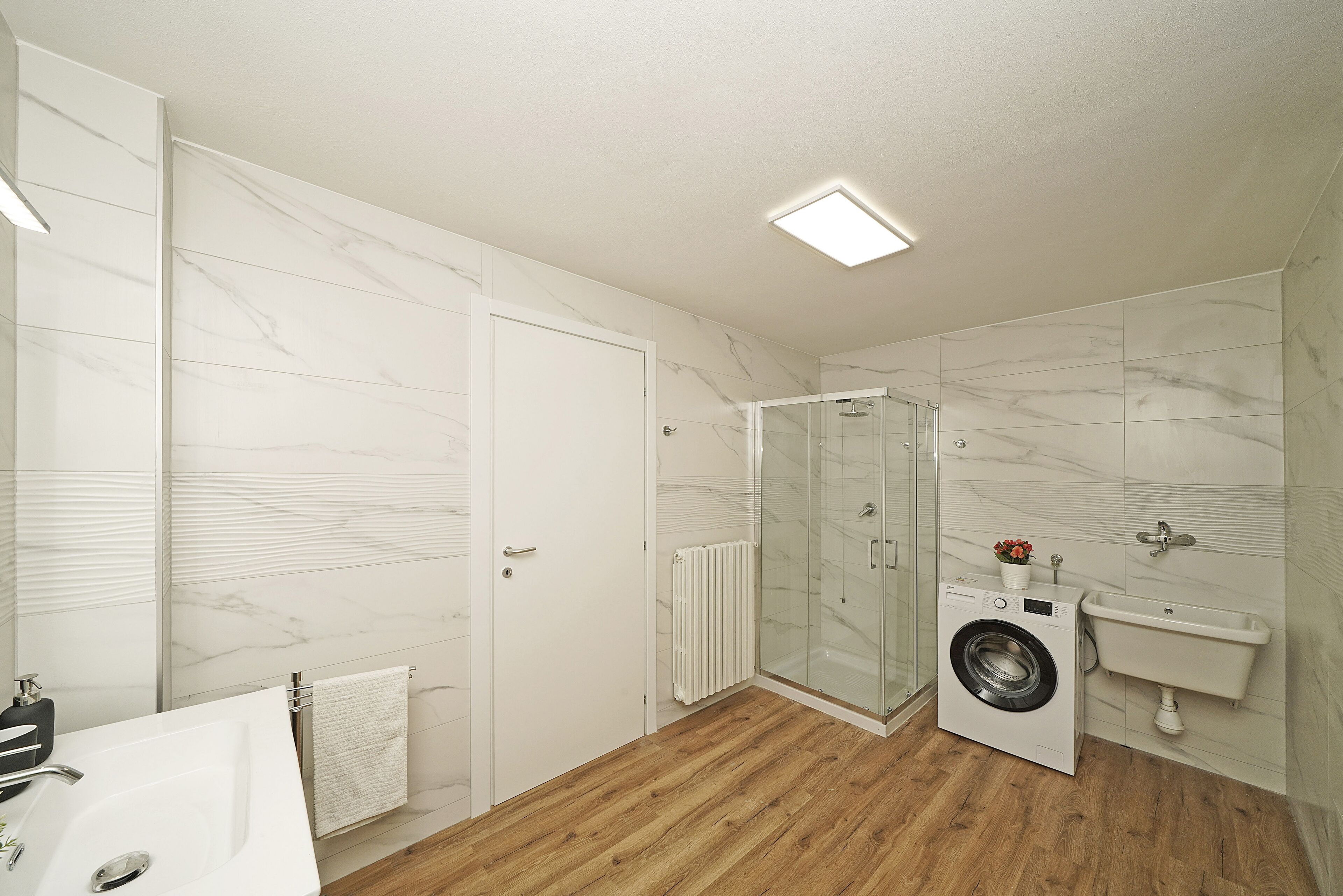 Un baño moderno con suelo de madera, cabina de ducha con mampara de vidrio, lavadora integrada y lavabo blanco.