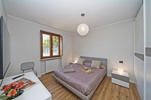 A bedroom with a bed, bedside table, lamp, window, and a desk with a remote control and a plant. - Casa Monte Cicogna PT-1 by Wonderful Italy (Moniga del Garda)
