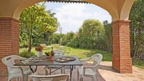 Une terrasse extérieure avec un pilier en briques, une table dressée avec des verres à vin et une bouteille, entourée de chaises blanches.