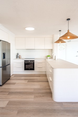A modern kitchen with white cabinetry, a built-in oven, a stainless steel refrigerator, and a large island with a sink.