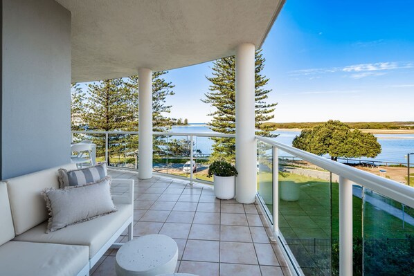 A balcony with a sofa and a round table, overlooking a body of water and greenery.
