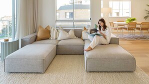 Un salon moderne avec un canapé d’angle, un coin repas comprenant une table et des chaises, et une grande fenêtre donnant sur un immeuble.