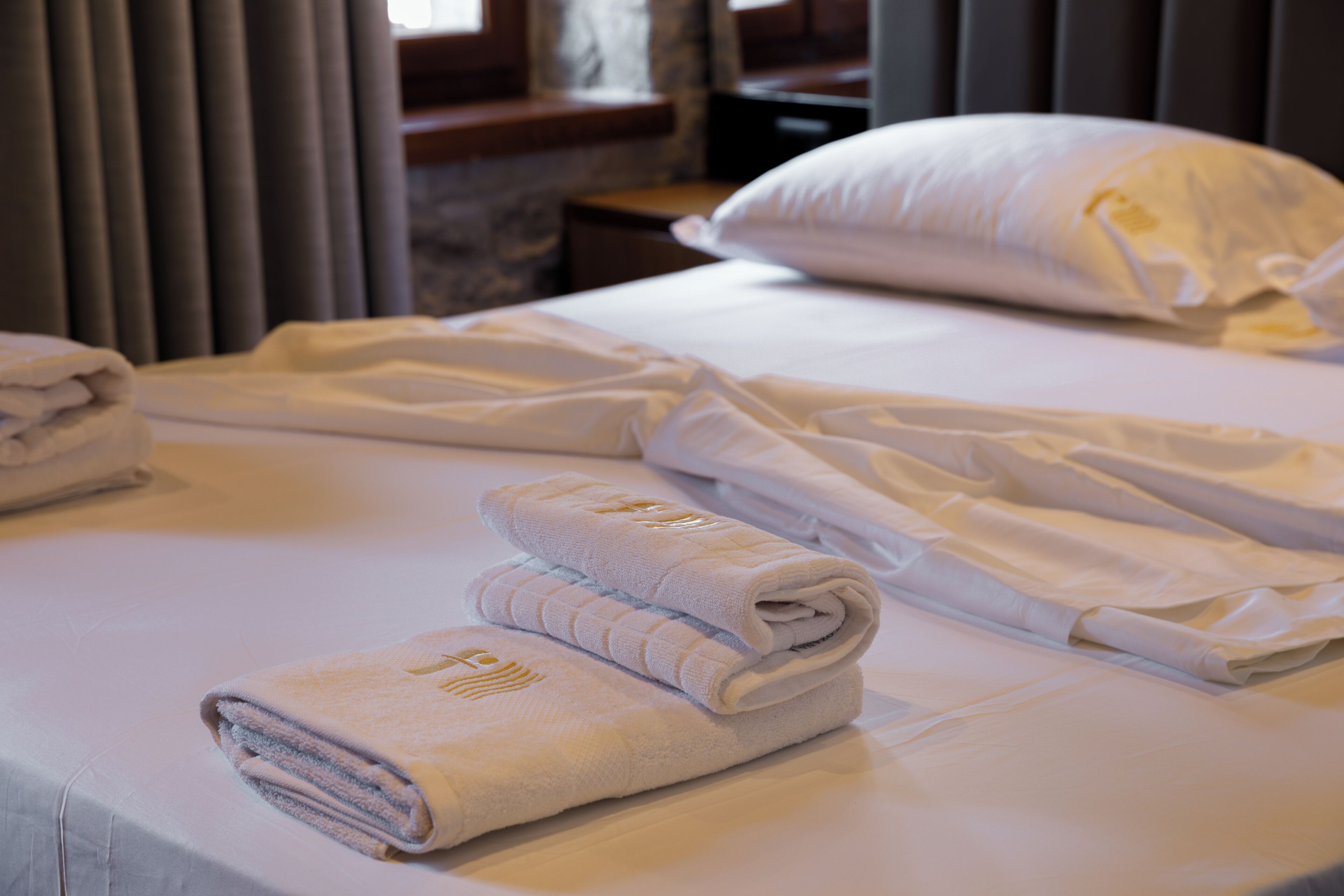 A neatly made bed with white linens and towels, a pillow, and a wooden nightstand.