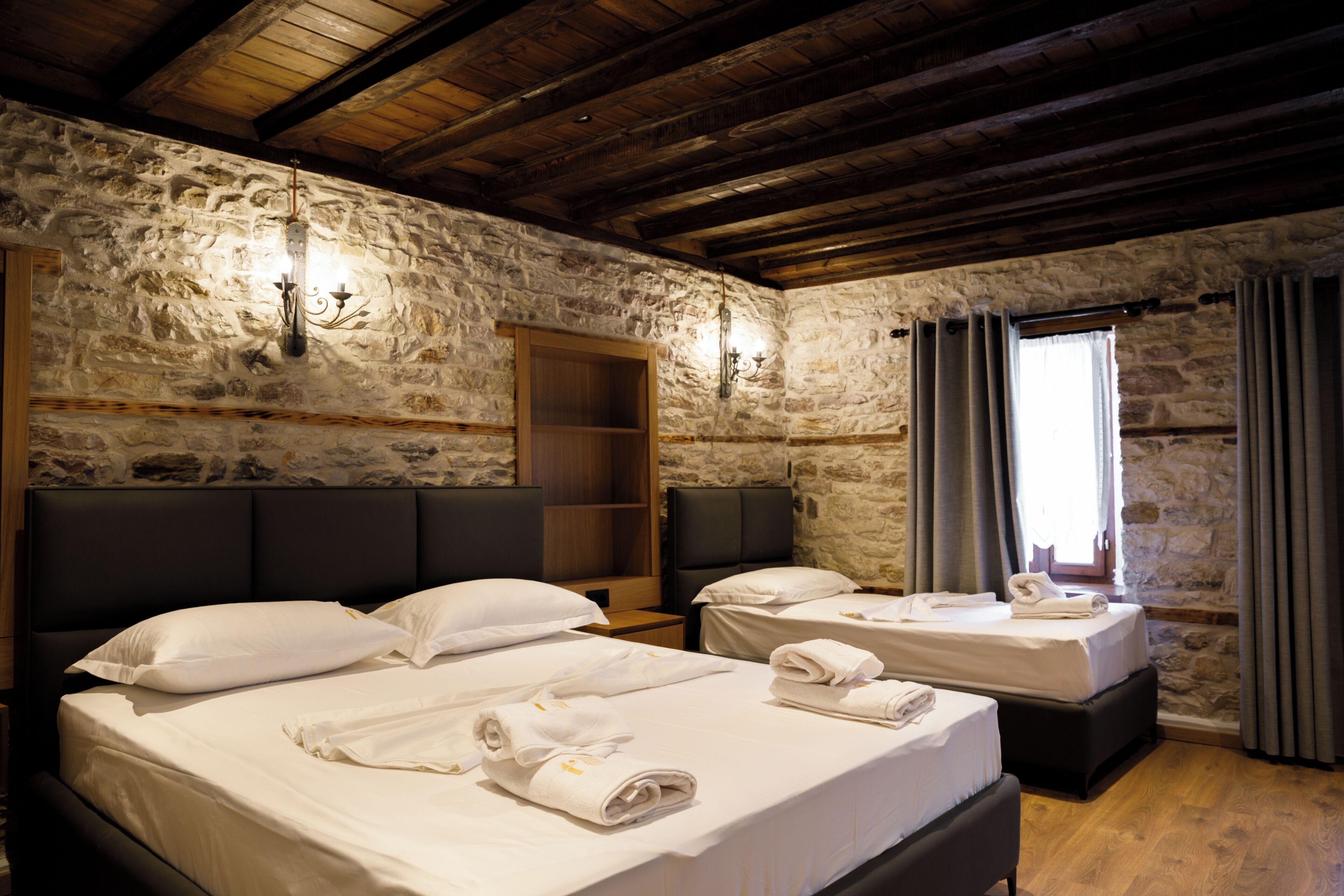 A room with two beds, stone walls, wooden beams, and a window with curtains.