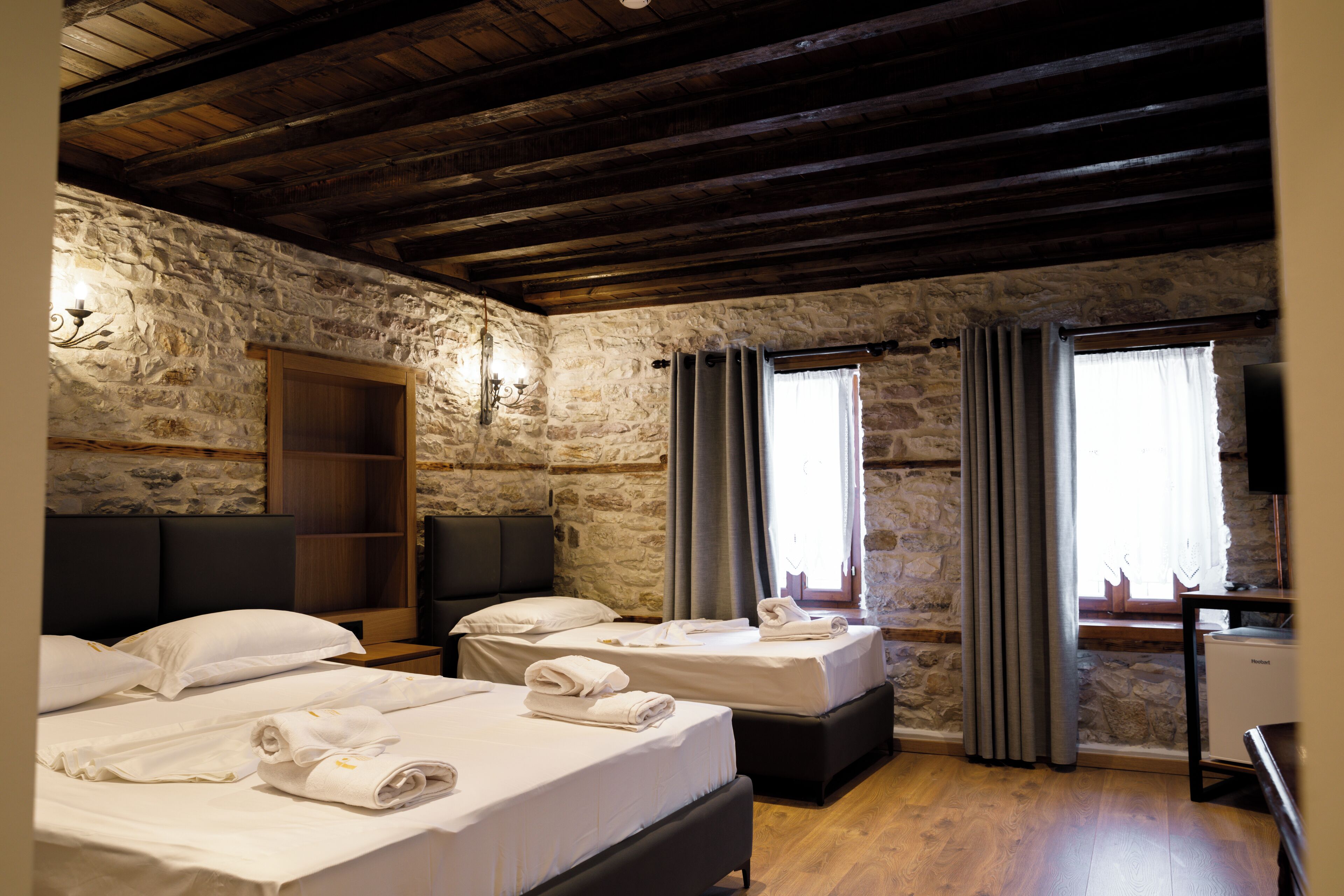 A room with two beds, stone walls, wooden beams, a desk, and a window with curtains.