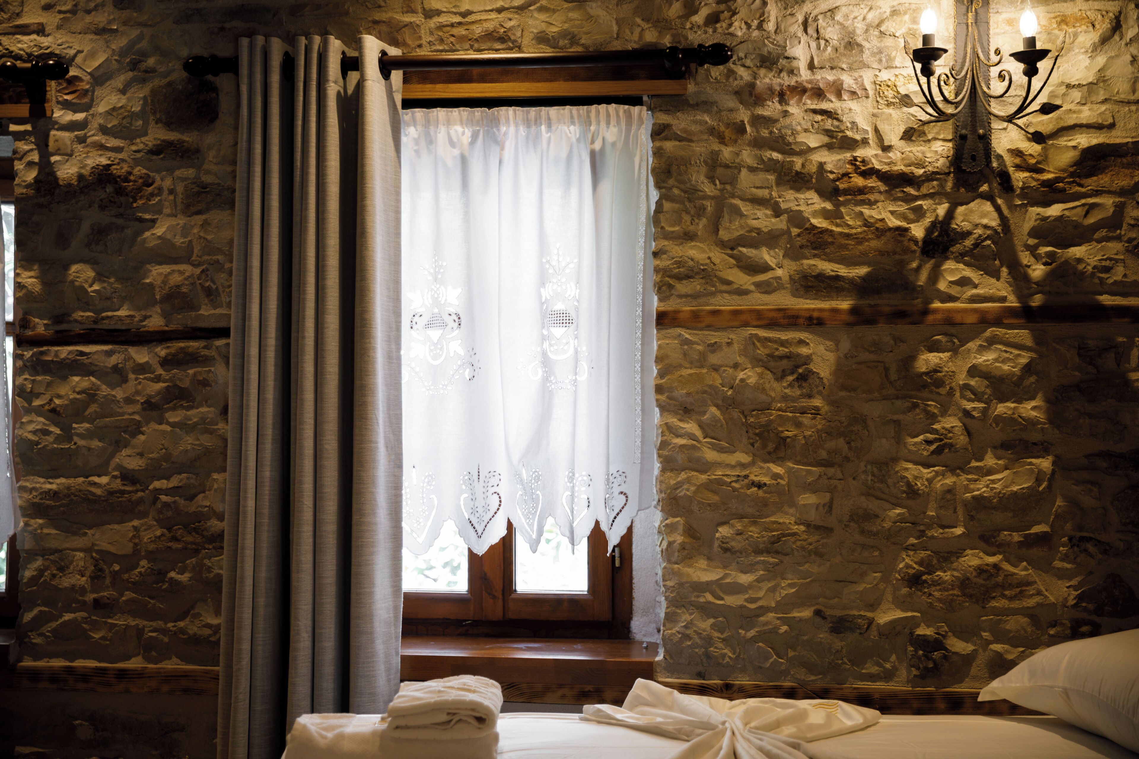 A room with a stone wall, a wooden window with curtains, and a bed with white linens.