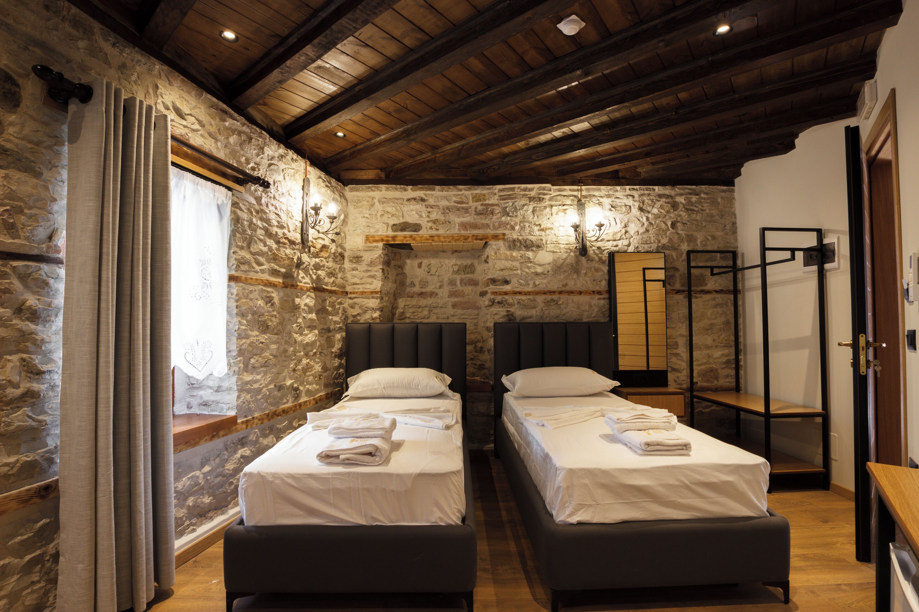 A room with two beds, stone walls, wooden beams, and a window with curtains.
