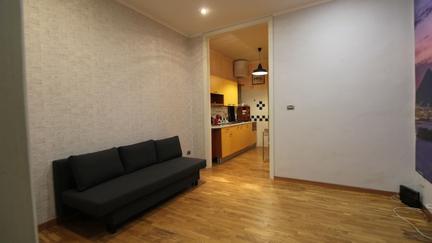 Een moderne woonkamer met een zwarte bank, houten vloeren en een keuken die zichtbaar is door een open doorgang.