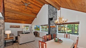 A spacious living area with a stone fireplace, wooden ceiling, and large windows offering a view of the outdoors.