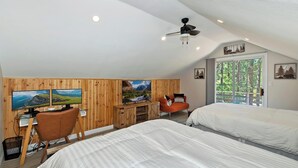 A bedroom with two beds, a desk with a computer, a chair, and a large window with a view of trees.