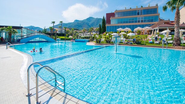 A large outdoor swimming pool with clear blue water, surrounded by palm trees and a modern building with multiple floors and large windows. - Residence Loano 2 Village Two-room Apartment 4 Pax Balcony (Loano)