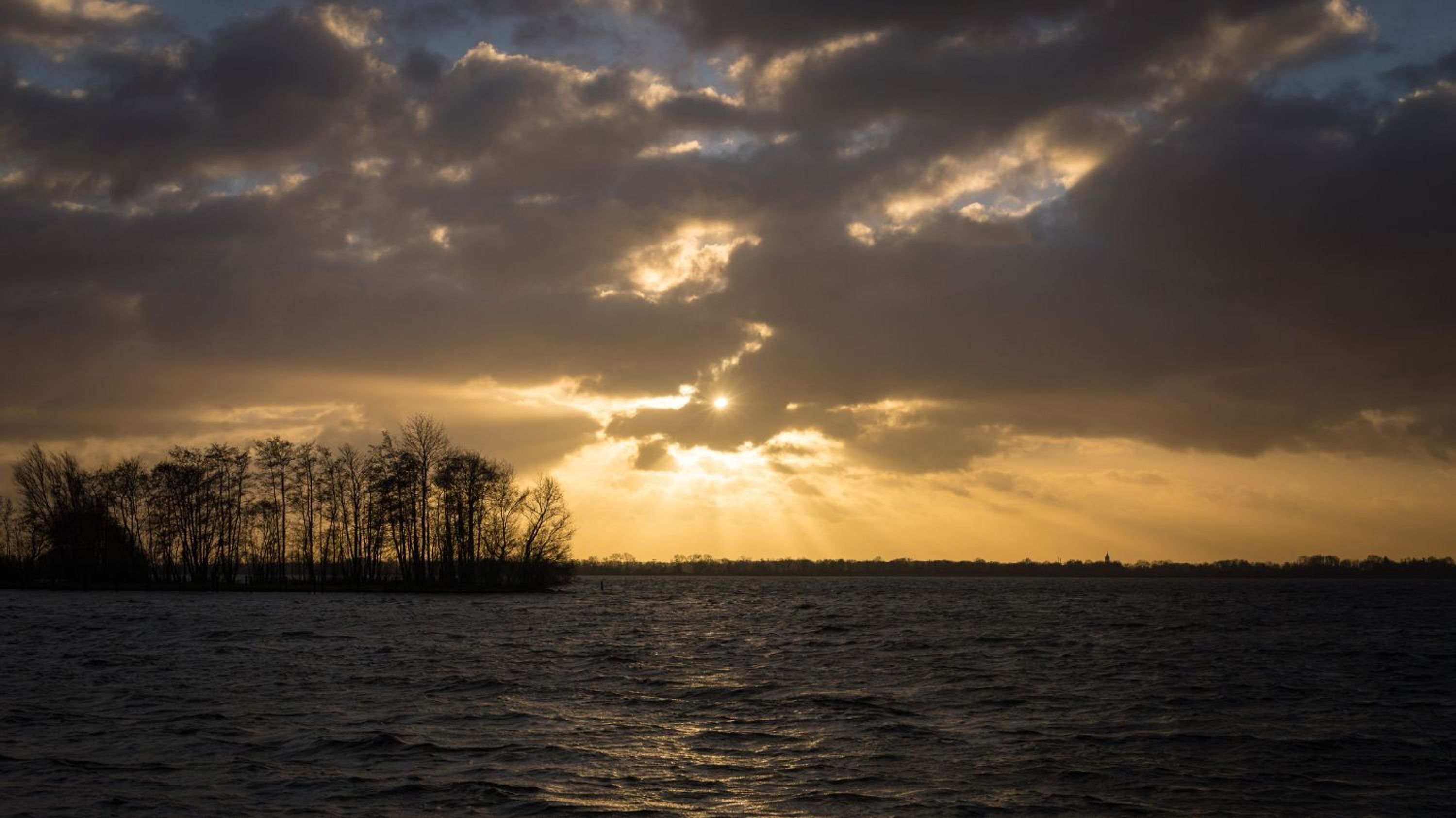 Un atardecer sobre un cuerpo de agua, con la silueta de árboles y una costa distante.