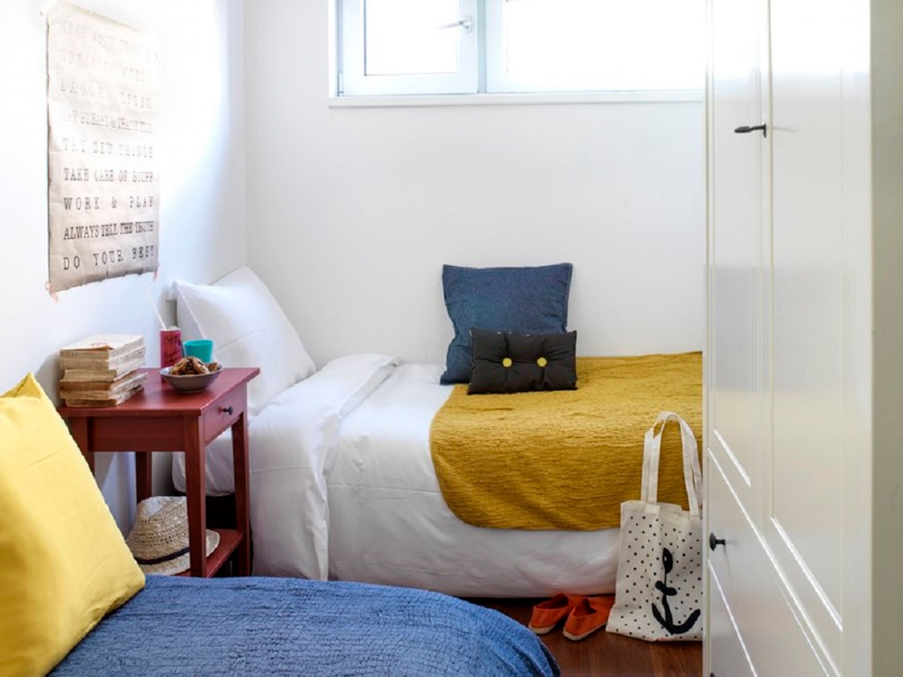 Une chambre avec deux lits, une fenêtre, un mur orné d’une affiche motivante, une petite table avec des livres et une tasse, et un sac blanc posé sur le sol.