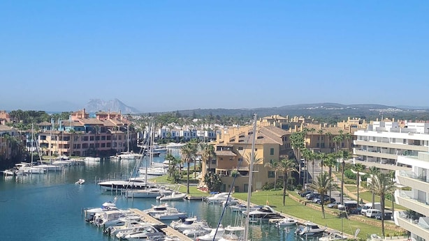 A marina with boats docked, surrounded by residential buildings and palm trees. - Waterside marina view 3 bedroom apartment with private pool in Puerto Sotogrande (San Roque)
