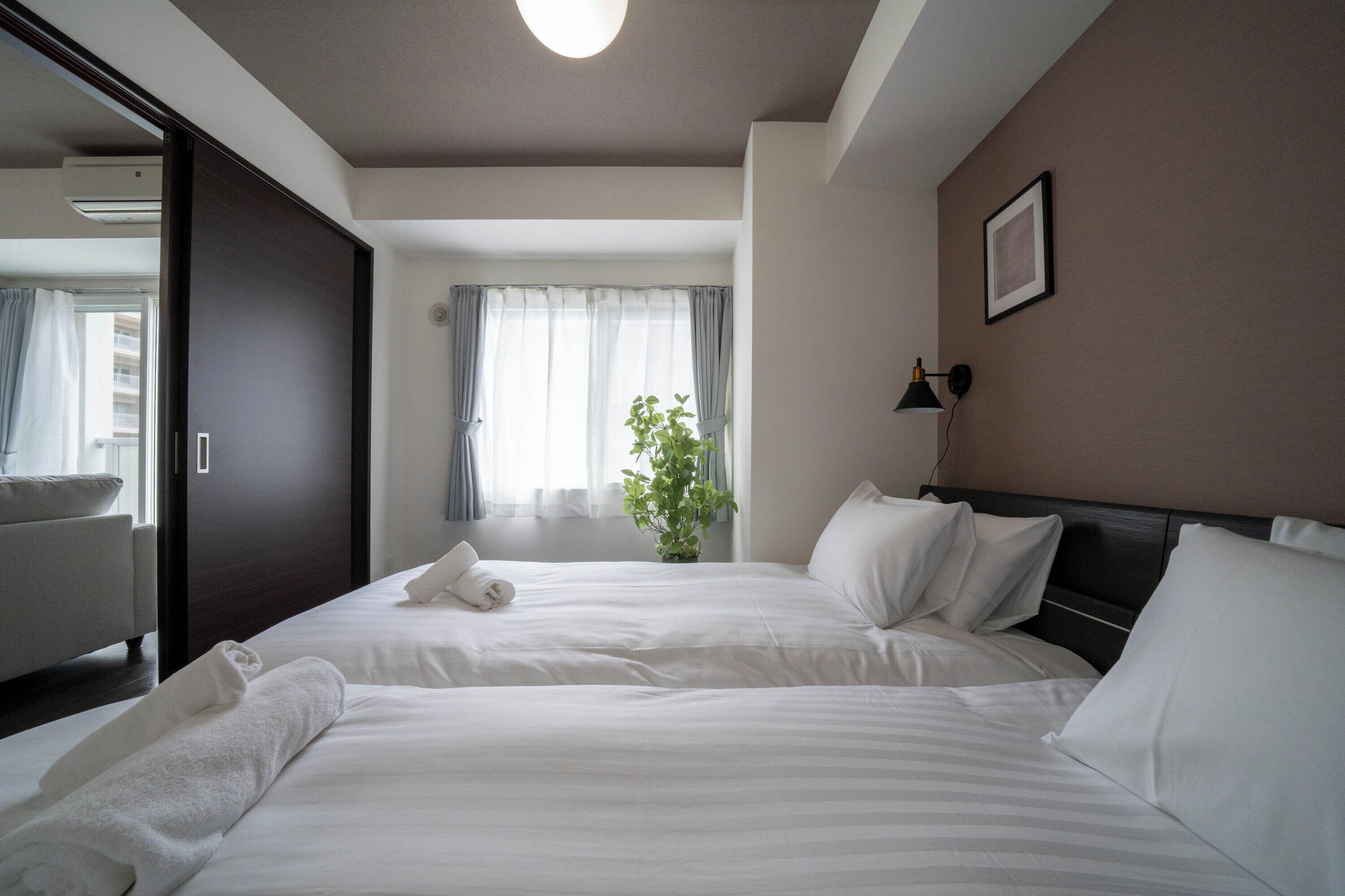 A hotel room with two beds, a large window with curtains, a potted plant, and a wall-mounted lamp.