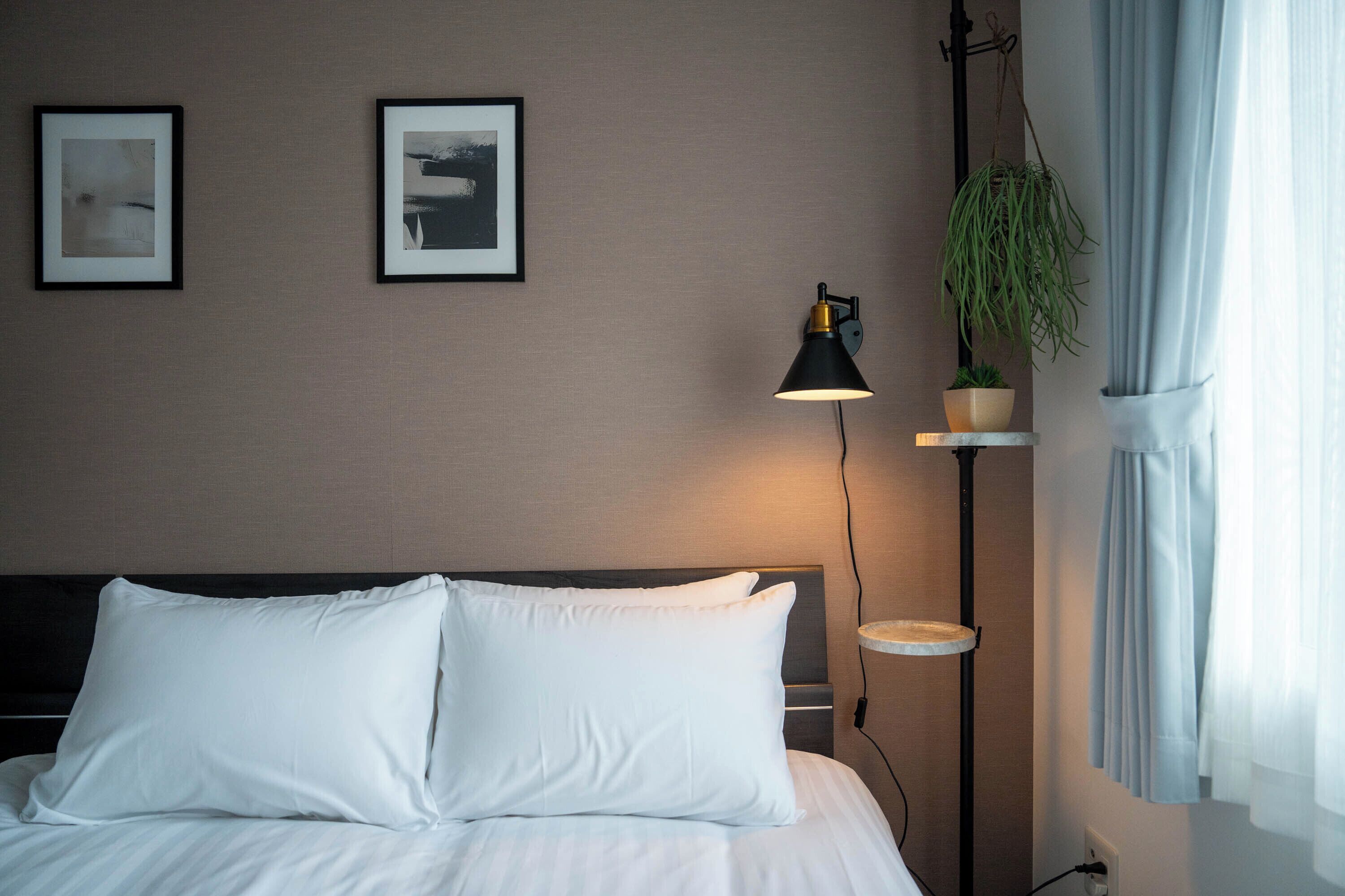 A neatly made bed with white linens, a black floor lamp, and two framed pictures on the wall.