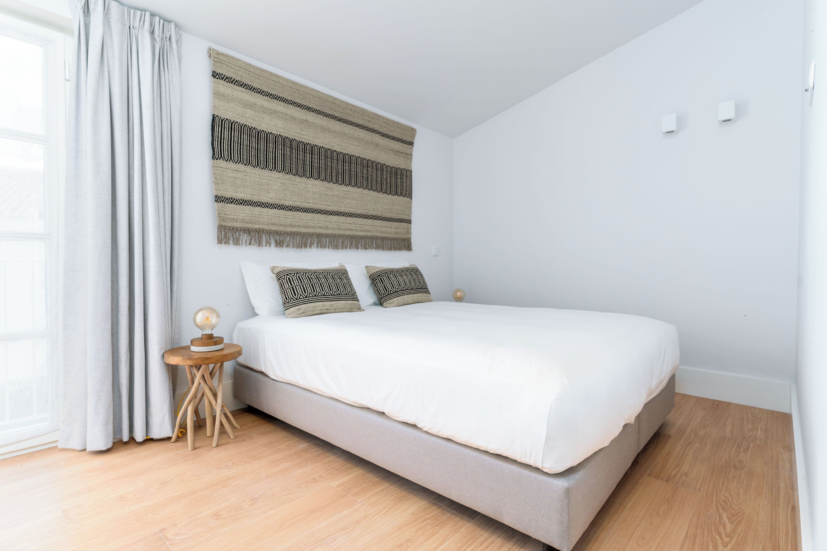 A bedroom with a bed, a small wooden nightstand, a wall-mounted tapestry, and a window with curtains.