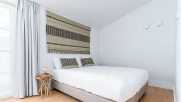 A bedroom with a bed, a small wooden nightstand, a wall-mounted tapestry, and a window with curtains.