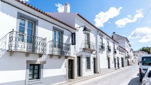 A white building with balconies, a cobblestone street, and a parked car. - Limehome Evora Rua de Machede 52 (Évora)
