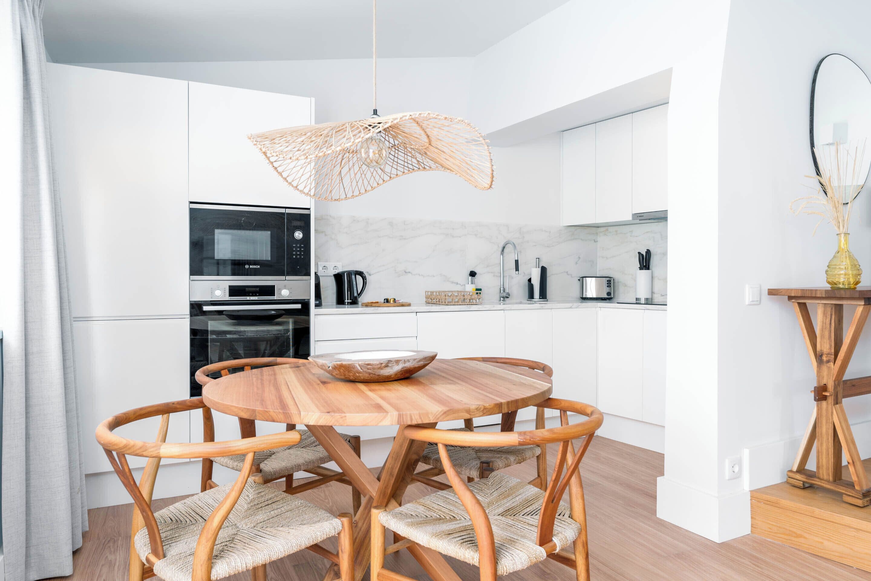 A modern kitchen with white cabinets, a wooden dining table with woven chairs, and a hanging light fixture.