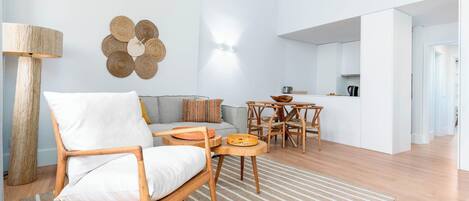 Una sala moderna con una mecedora de madera, un sofá, una mesa de comedor con sillas y una zona de cocina al fondo.