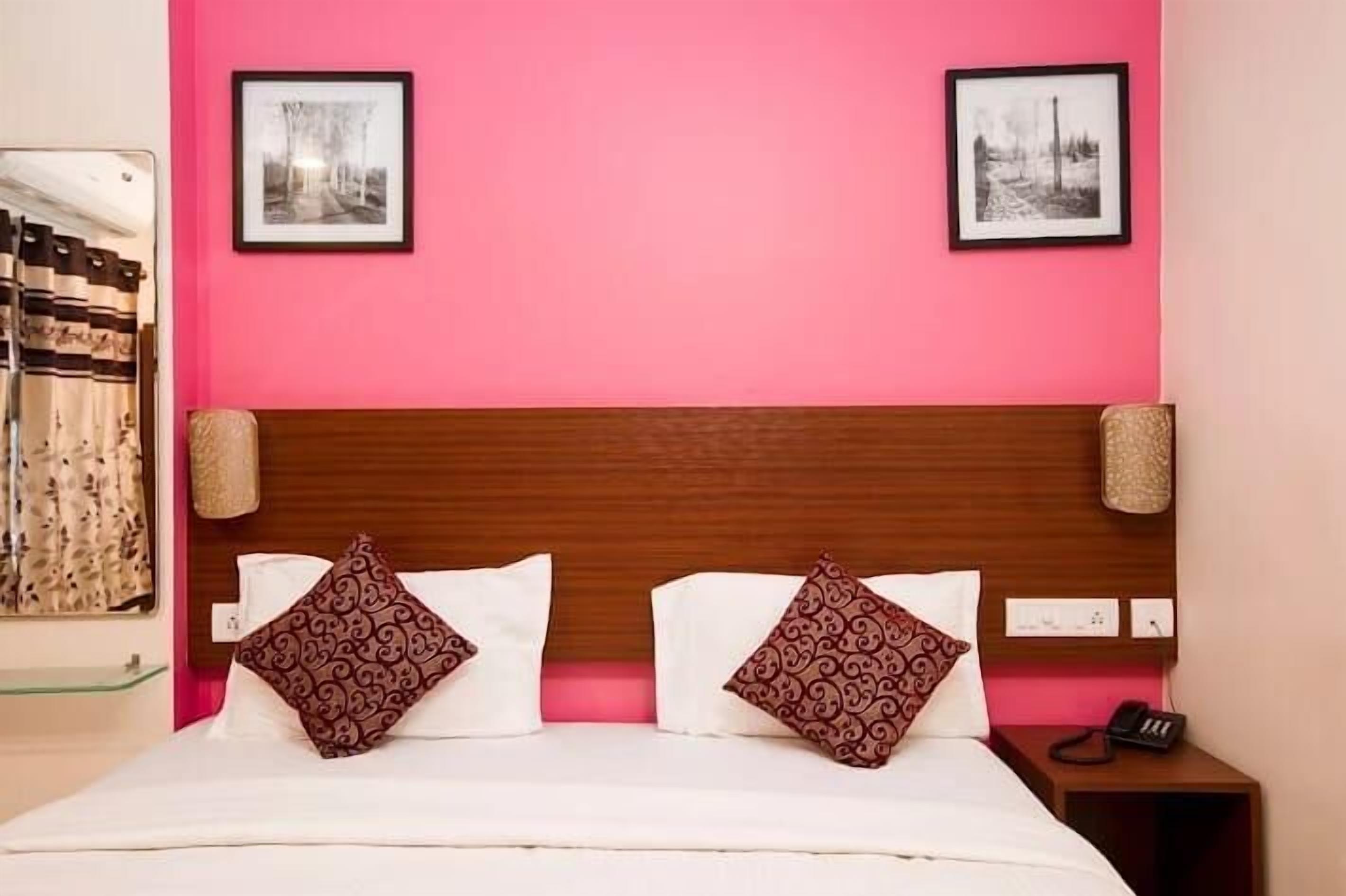 Habitación de hotel con pared rosa, cabecera de madera, dos camas con almohadas estampadas, un espejo y dos cuadros enmarcados.