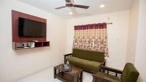 A living room with a flat-screen TV mounted on the wall, a ceiling fan, a green sofa, a wooden coffee table, and a patterned curtain. - Terra Hotels T nagar (Chennai)