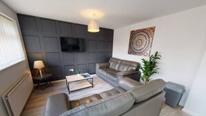 A modern living room with a grey sofa, a coffee table, a lamp, and a TV mounted on a dark wall. - Book Direct 10% Off > Flexley House 4 Br Workstays (Middlesbrough)