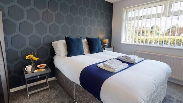 A hotel room with a large bed, a nightstand with a lamp, a window with blinds, and a wall with a geometric pattern. - Book Direct 10% Off > Flexley House 4 Br Workstays (Middlesbrough)