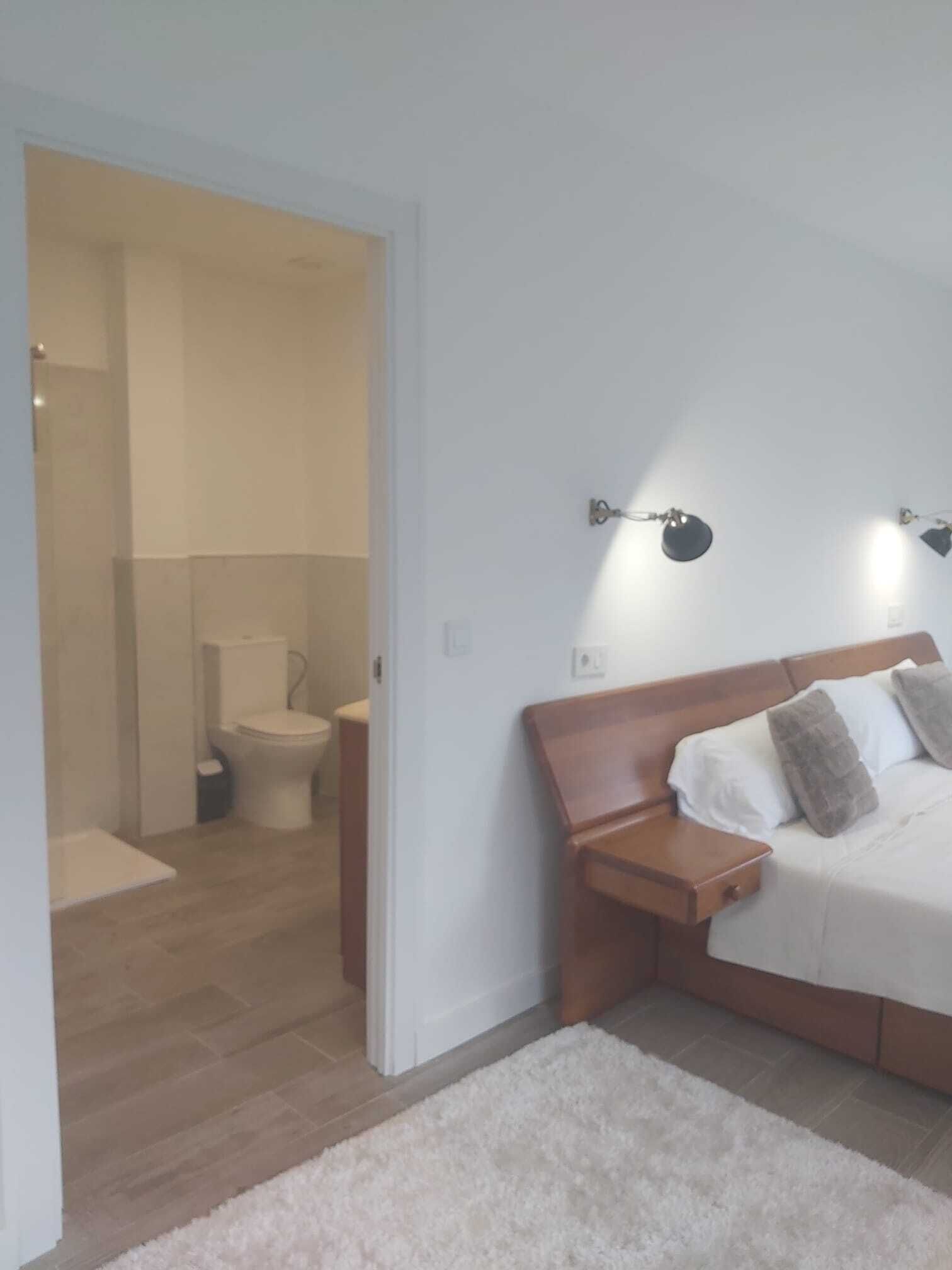 Une chambre avec une tête de lit en bois, un lit recouvert de linge de lit blanc, une table de chevet et un tapis blanc. On aperçoit, au fond, une salle de bain avec des toilettes et un espace douche.