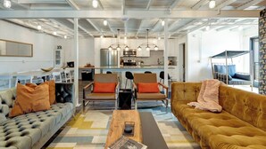 Ein modernes Wohnzimmer mit einer Couch, Sesseln und einem Couchtisch. Im Hintergrund ist eine Küche mit Essbereich zu sehen.