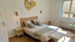 A bedroom with a wooden floor, a bed with a white bedspread and multiple pillows, a bench at the foot of the bed, a small wooden nightstand, a wall-mounted light fixture, and a window with a view of the outside. - 2 Bedroom Private vacation home in Amboise (Amboise)