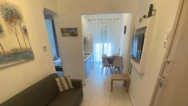 A narrow hallway with a dark-colored sofa on the left, a dining area with a table and chairs in the center, and a kitchen visible through an open door in the background. - Cozy Apartment With two Bedrooms With a sea View (Thermaikos)