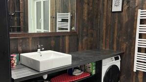 A modern bathroom with a wooden wall, a white sink, a mirror, a towel rack, a washing machine, and a small stool. - 2 self-contained country homes with heated indoor swimming pool 8-12p (Aix-en-Issart)