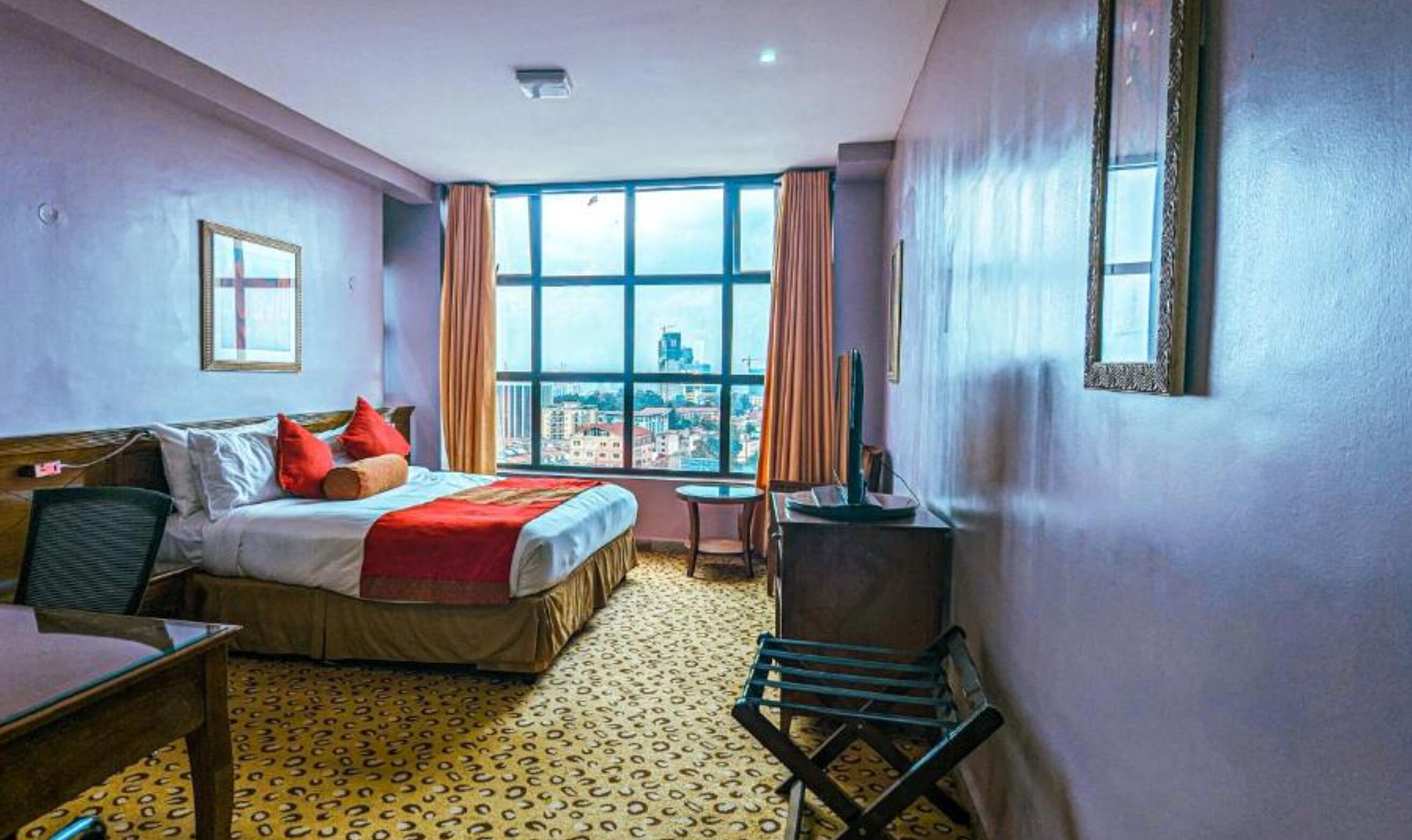 A hotel room with a bed, a desk, a chair, a TV, and a window with a city view.