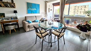 A modern living room with a glass dining table, a sofa, and a balcony with a view of buildings.