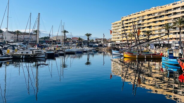 A marina with boats docked, a large hotel building, and clear skies.