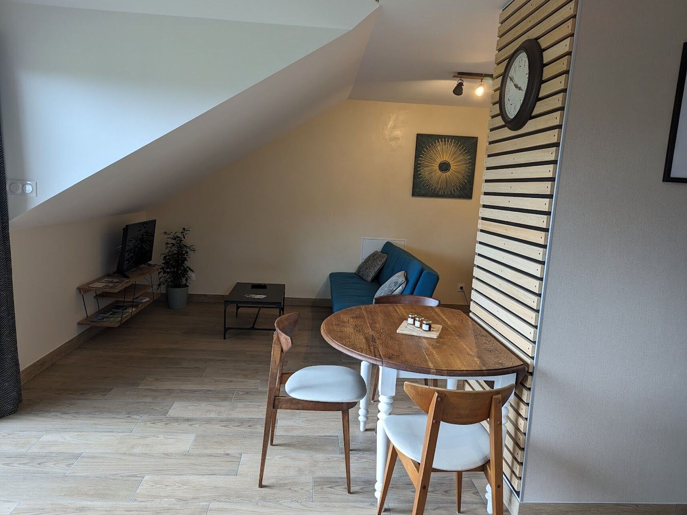 A loft-style living space with a dining area, a blue sofa, a wooden wall panel, and a clock.